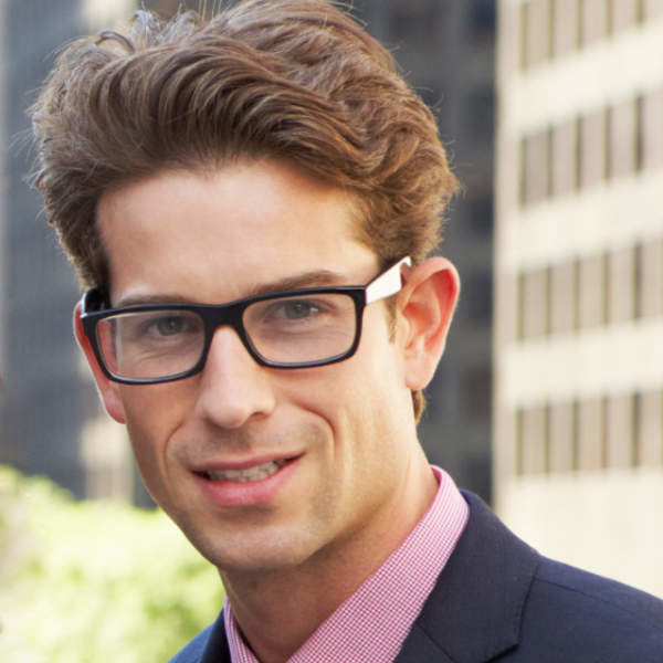 Young man with glasses smiling outdoors in an urban setting.