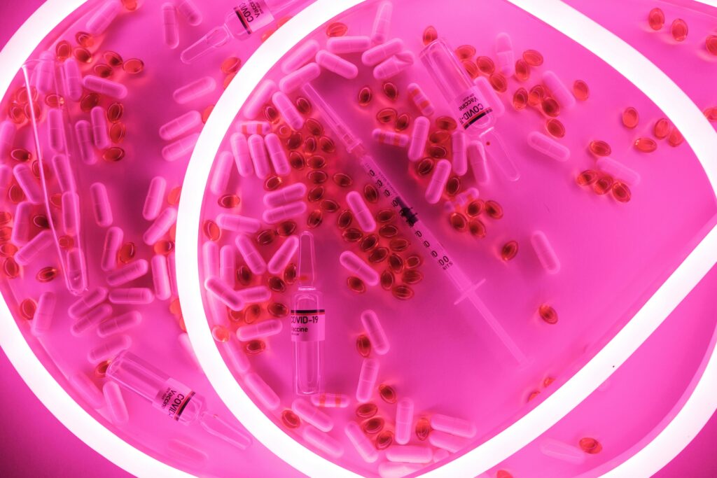 A vibrant neon pink composition of Covid-19 vaccine vials, capsules, and syringe viewed from above.